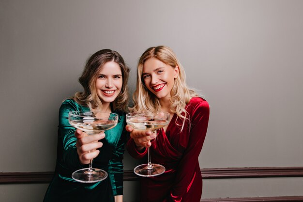 Good-looking female models posing with wineglasses on gray wall. Indoor shot of charming ladies in velvet dressees celebrating something.