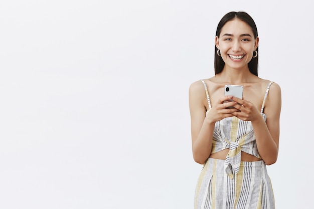 Bella femmina dai capelli scuri diabolica e felice con la talpa sotto il labbro, sorridendo con gioia mentre guarda e tiene lo smartphone, controllando la nuova app.