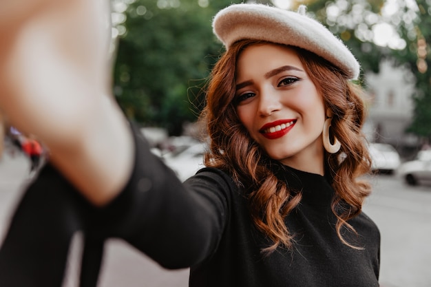 Good-looking french girl with ginger hair posing in november day. Outdoor shot of elegant caucasian lady with red lips making selfie on street wall.