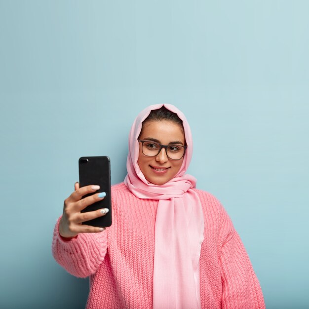 Good looking girl with gentle smile, looks gladfully at device screen, wears rectangular spectacles, silk veil, extends hand for making nice shot, has manicure, dressed in pink jumper, isolated on blue