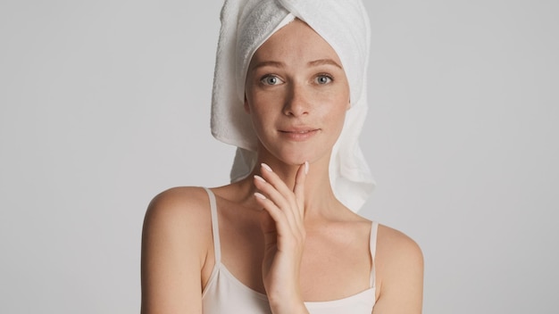 Good looking girl with smooth healthy skin wearing towel on head looking straightly in camera over white background