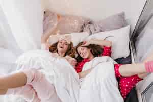 無料写真 ベッドに横になって幸せを表現するイケメン。週末にポーズをとる明るいパジャマで笑っている姉妹の屋内写真。