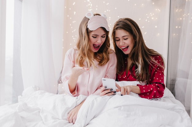 Good-looking girls in stylish pajamas texting phone message. Indoor portrait of glamorous young ladies sitting in bed together and looking at smartphone screen.