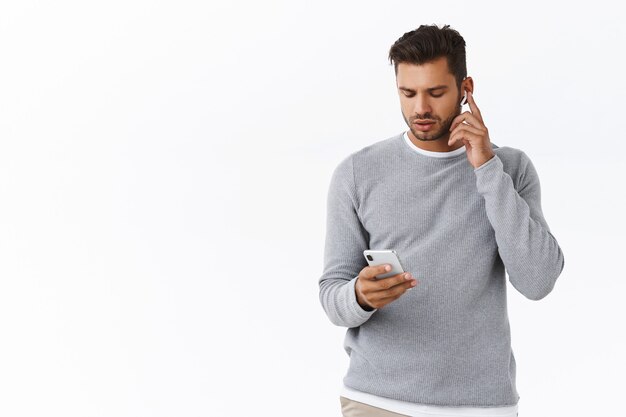 Good-looking guy getting ready leave office, put on wireless headphones, increase volume touch earphone, holding smartphone, looking mobile screen as changing earbuds settings, white wall