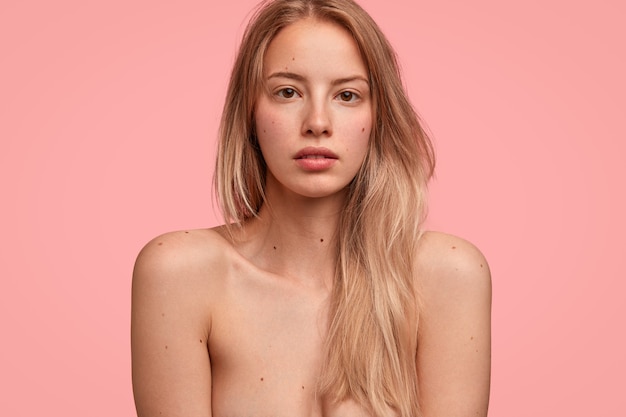 格好良い半裸の女性はスリムな完璧な体を示し、長い明るい髪を持ち、ピンクの壁に隔離されたカメラで直接真剣な表情で見えます