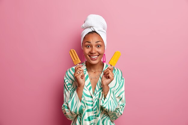 Good looking healthy young woman holds two popsicles on sticks, has happy mood, wears casual domestic clothes, enjoys last days of summer, poses 