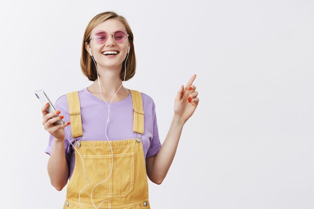 Good-looking joyful woman dancing with closed eyes carefree, holding smartphone, listening music in earphones