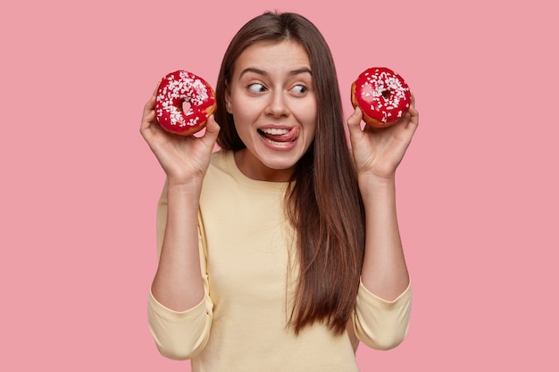 good looking lady licks lips with tongue, eats donuts, looks positively aside