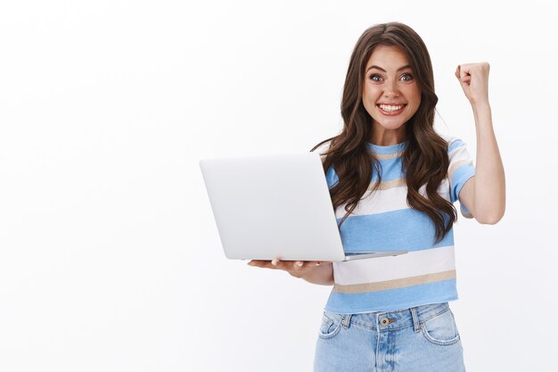 Красивая счастливая успешная европейская женщина держит ноутбук, поднимает кулак, торжествуя, радостно улыбается, получает одобрение, получила повышение, выигрывает онлайн-игру, празднует победу