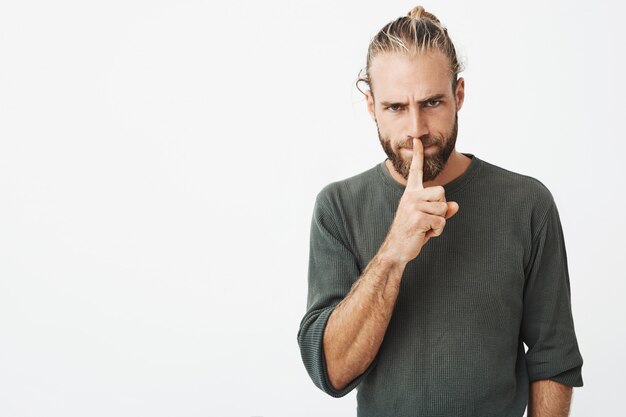 Good-looking mature guy with stylish hairdo and beard holding index finger at lips, asking for silent