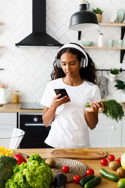 Good-looking mulatto woman is looking on the smartphone and greenery, in big wireless headphones, near the table with fresh vegetables