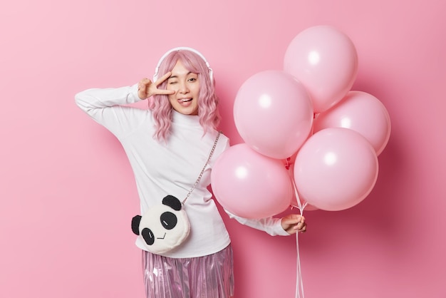 Good looking pink haired Asian girl makes peace gesture over eye sticks out tongue foolishes around on party dressed festively holds bunch of inflated helium balloons poses for making photo