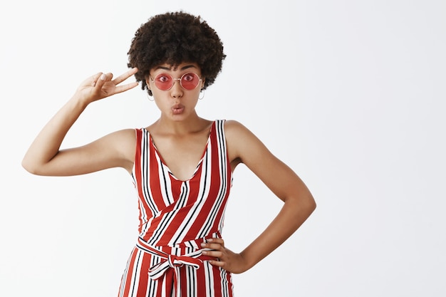 Free photo good-looking and playful dark-skinned girl with curly hairstyle, holding hand on hip, showing peace sign and folding lips joyfully, standing