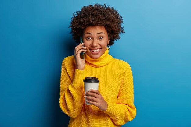 Good looking positive Afro American woman has telephone conversation, holds mobile phone near ear