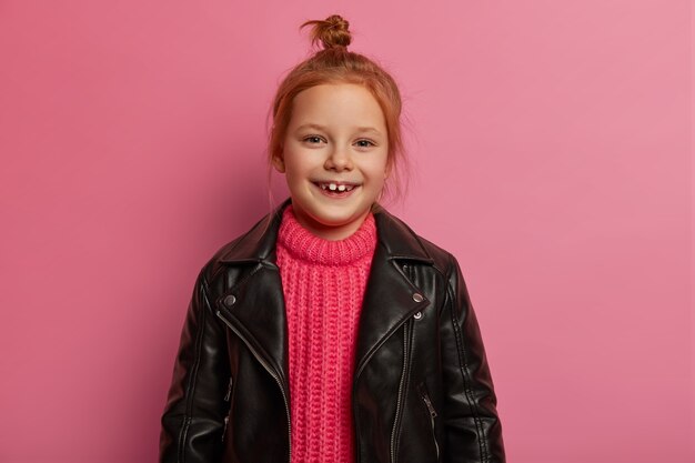 Good looking positive little kid has ginger hair combed in knot, wears rosy knitted sweater and leather black jacket, feels playful, glad after successful shopping day, stands against pink wall.