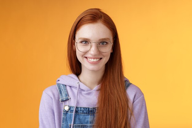 かっこいい赤毛の女性プログラマーメガネパーカー笑顔満足した外観プロの目的成功立っている自信を持ってニヤリと喜んでカメラがカジュアルにオレンジ色の背景を話している プレミアム写真