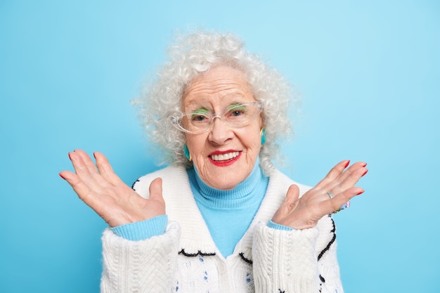 잘 생긴 은퇴 한 여성이 손바닥을 펴고 부드럽게 미소를 지으며 긍정적으로 투명한 안경을 쓰고 스웨터를 입는다.