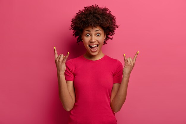 Симпатичная нахальная женщина демонстрирует рок-н-ролльный жест, веселится, слушает любимую музыку, радостно восклицает, имеет эмоциональное выражение, носит повседневную футболку, изолированную на розовой стене. Знак хэви-метала