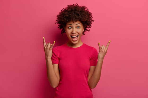 Good looking sassy woman shows rock n roll gesture, has fun, listens favorite music, exclaims wih joy, has emotional expression, wears casual t shirt, isolated on pink wall. Heavy metal sign