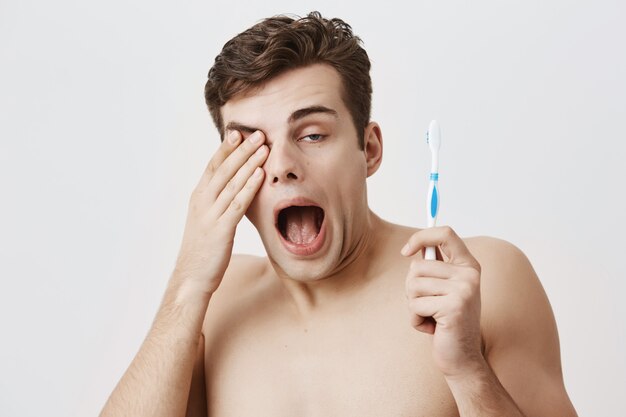 Good-looking sleepy male student with trendy hairstyle, woke up early in the morning, getting ready for work or university. Muscular guy yawning, holding toothbrush in his hand, rubbing eye.