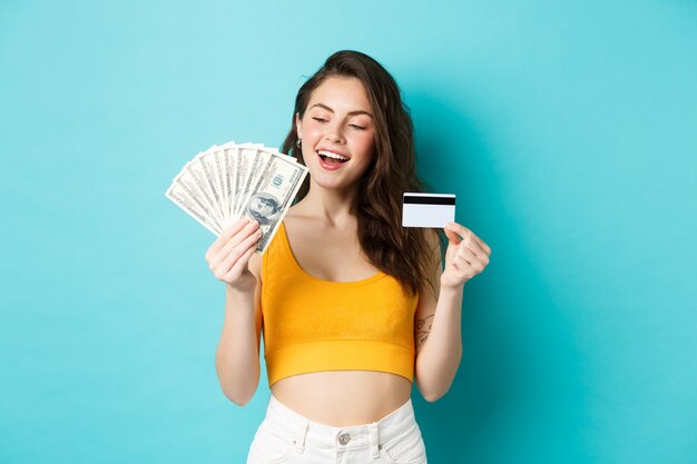 クレジットカードを持って、青い背景に立って、ドル紙幣のお金に満足し、決心している見栄えの良い笑顔の女の子。