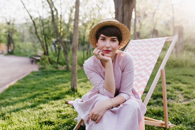 ピクニックを待っているリクライニングチェアに座っている昔ながらの帽子をかぶった格好良いスタイリッシュな若い女性