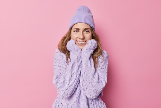 Good looking tender millennial woman keeps hands under chin smiles toothily wears hat and sweater looks happily listens pleasant words isolated over pink background. Human emotions face expressions