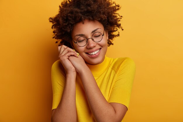 Good looking tender young female model leans at hands, smiles tenderly, closes eyes and imagines something wonderful or pleasant, being touched, wears spectacles and yellow outfit, has romantic mood
