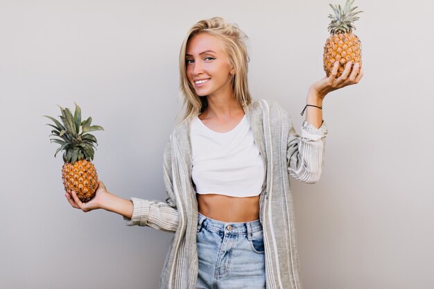 Good-looking trendy woman with pineapples smiling to camera. blonde woman in casual attire.