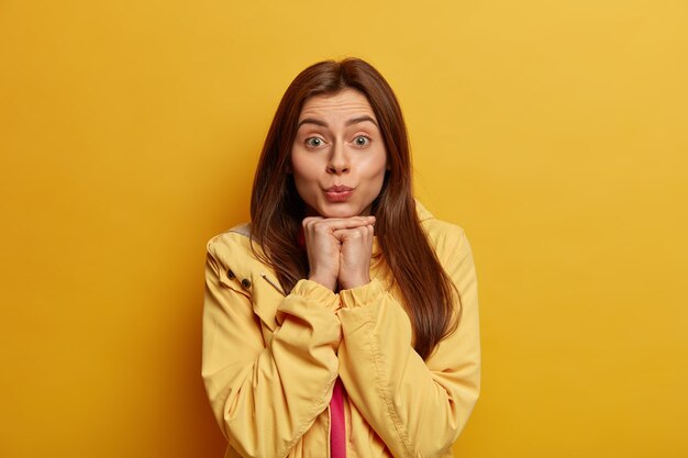 Good looking woman keeps lips folded, wants to kiss someone, looks directly at camera, holds hands under chin, wears yellow anorak