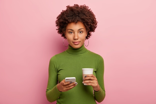 Good looking woman looks straigly at camera with confident expression, holds mobile phone, views photos in social networks, drinks takeout coffee