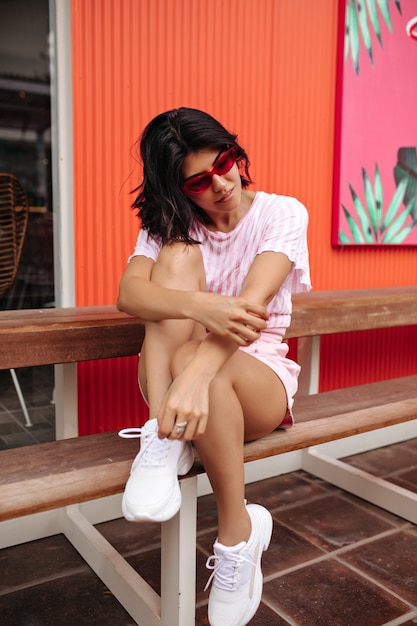 Good-looking woman in white sneakers sitting on wooden bench. Outdoor shot of refined european woman in summer attire.