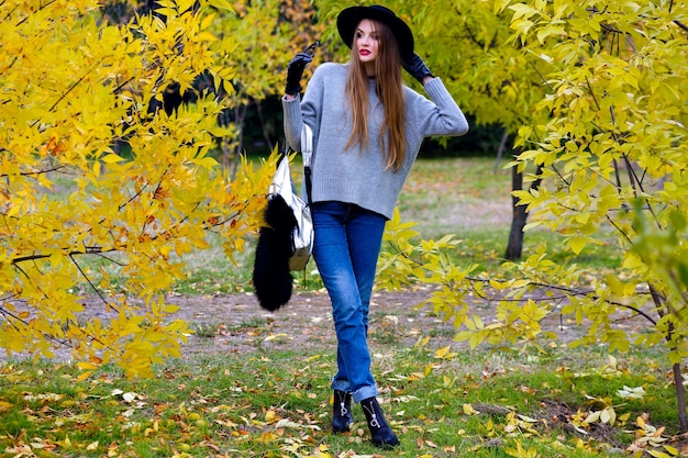 Good-looking woman with long hair wears jeans and gloves standing in confident pose on nature background. Outdoor photo of pretty female model in trendy gray sweater walking in park in autumn day.