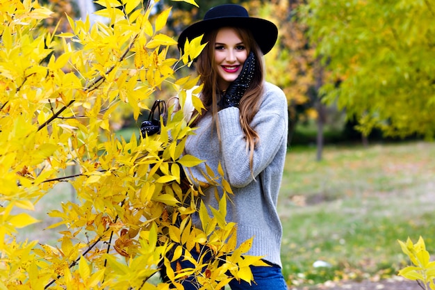 Good-looking woman with long hair wears jeans and gloves standing in confident pose on nature background. Outdoor photo of pretty female model in trendy gray sweater walking in park in autumn day.