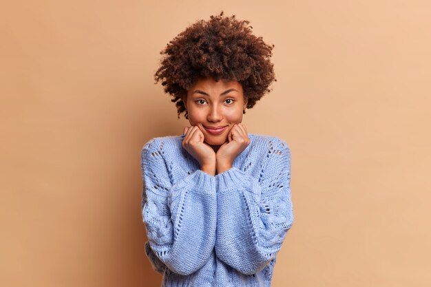 Good looking woman with natural curly hair keeps hands under chin dressed in blue jumper looks directly at front stands confident and beautiful stands against brown wall