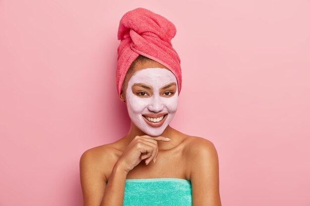 Красивая женщина с довольным выражением лица, нежно касается подбородка, носит очищающую глиняную маску на лице, обернута полотенцем на голове, наслаждается косметическими процедурами дома, изолирована на розовой стене