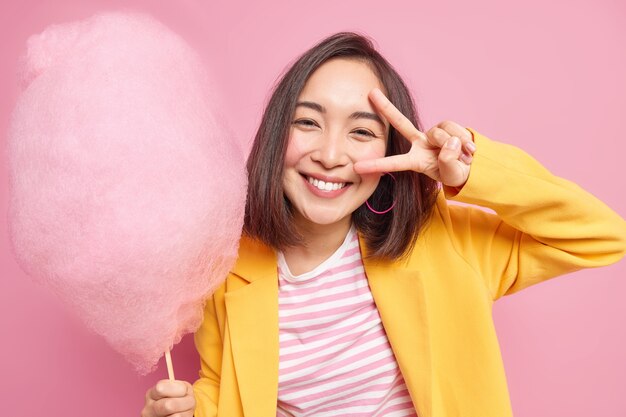 Good looking young Asian woman smiles positively makes victory gesture over eye has upbeat mood holds delicious candy floss wears yellow jacket has sweet tooth poses against pink wall.