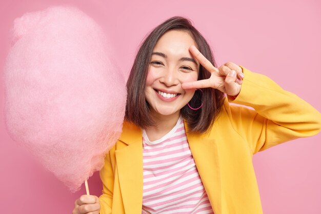 Симпатичная молодая азиатская женщина позитивно улыбается, делает победный жест над глазами, имеет оптимистичное настроение, держит вкусную сахарную вату, носит желтую куртку, принимает позы для сладкоежек на фоне розовой стены.