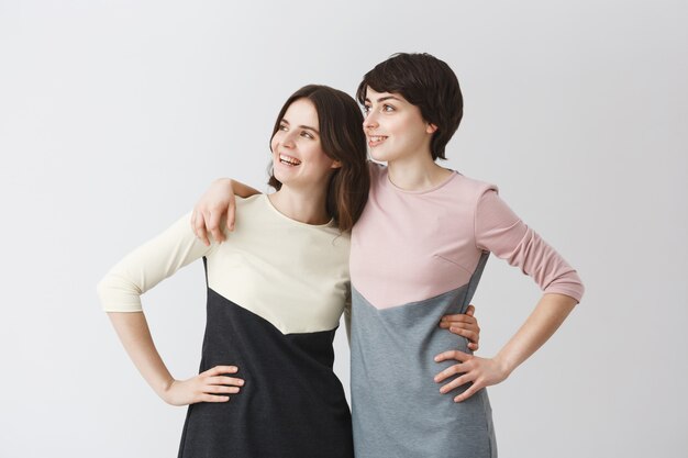 Good-looking young cheerful lesbian girl with dark short hair hugging her long-haired beautiful girlfriend, looking aside, posing for photo on a queer party.