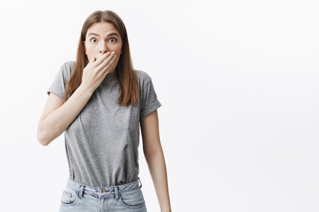 Good-looking young european dark-haired student girl with medium hair with frightened face expression, clothing mouth with hand, being shocked after saying something she not