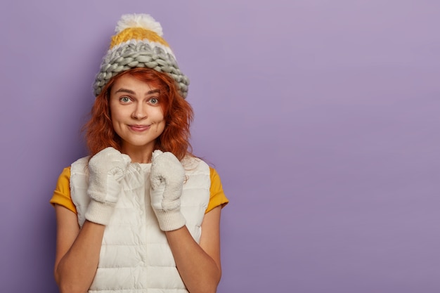 Good looking young female raises clenched fists, wears vest, t shirt, white gloves, headgear on head, looks with intriguing gaze at camera, isolated on purple wall