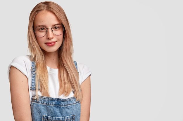 Foto gratuita bella giovane donna con un'espressione del viso soddisfatta, ha i capelli lunghi, la pelle sana, indossa occhiali rotondi e salopette di jeans