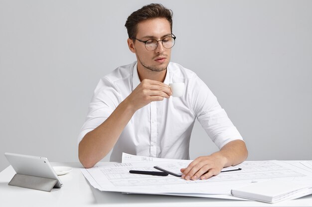 Good looking young man looks at cup as drinks hot coffee or tea, works at future project, develops new strategy, uses modrn device, sits indoors on work place. People, occupation, job concept