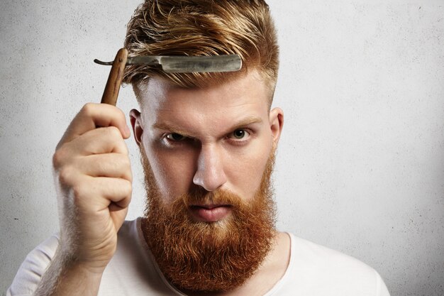 Good-looking young man with red beard holding barbershop accessory. Caucasian barber demonstrating sharp blade of his old-fashioned straight razor, determined to shave clients. 