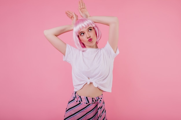 Good-looking young woman in casual stylish t-shirt fooling around . Indoor portrait of romantic lady in pink peruke making faces on bright wall