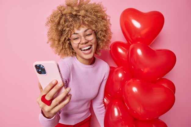 Good looking young woman makes selfie with smartphone smiles broadly wears transparent glasses holds bunch of inflated heart balloos isolated over pink background people and celebration concept