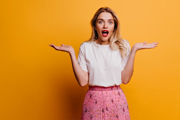 Good-looking young woman in white t-shirt posing with hands up. Indoor shot of emotional lady isolated on bright wall.