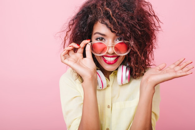 Красивая молодая женщина с коричневой кожей держит розовые солнцезащитные очки и позирует с удивленной улыбкой. Крытый портрет эмоциональной африканской женской модели в элегантной желтой одежде.