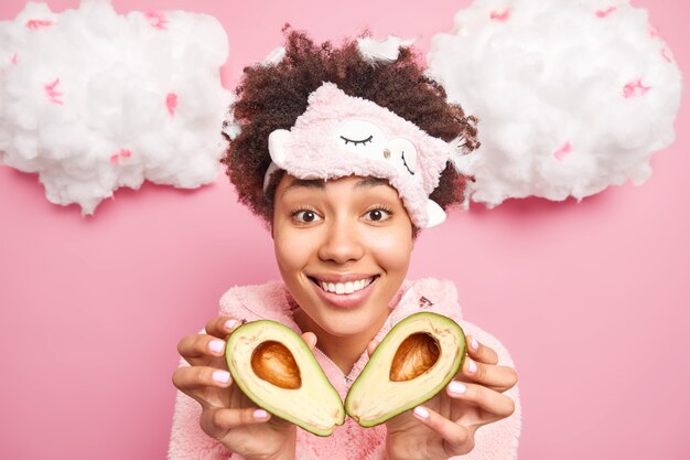 Good looking young woman with curly hair holds halves of avocado going to make natural cosmetic product to care about skin wears sleepmask poses against pink wall has feathers in hair