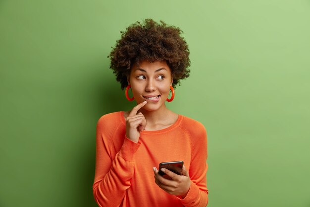 巻き毛の格好良い若い女性は、現代のスマートフォンを保持します緑の壁に隔離されたカジュアルなオレンジ色のセーターに身を包んだ新しいアプリケーションを使用